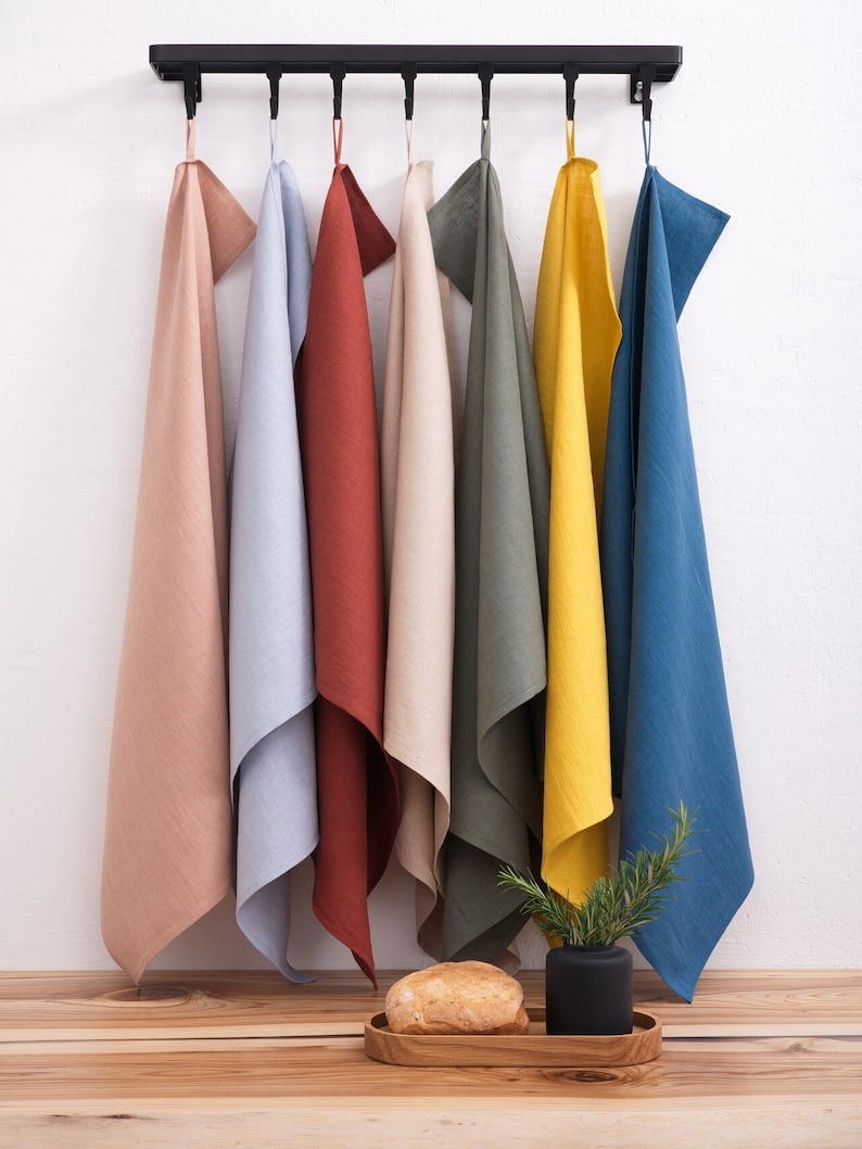 Colorful linen towels in shades of beige, blue, rust, olive, and yellow, hanging on a black metal rack against a white wall with a wooden surface below, where a loaf of bread and a potted plant are placed.