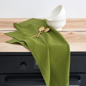 A vibrant green linen towel casually draped over a wooden kitchen countertop, accompanied by a stack of white textured bowls and a pair of wooden spoons, against a white wall.