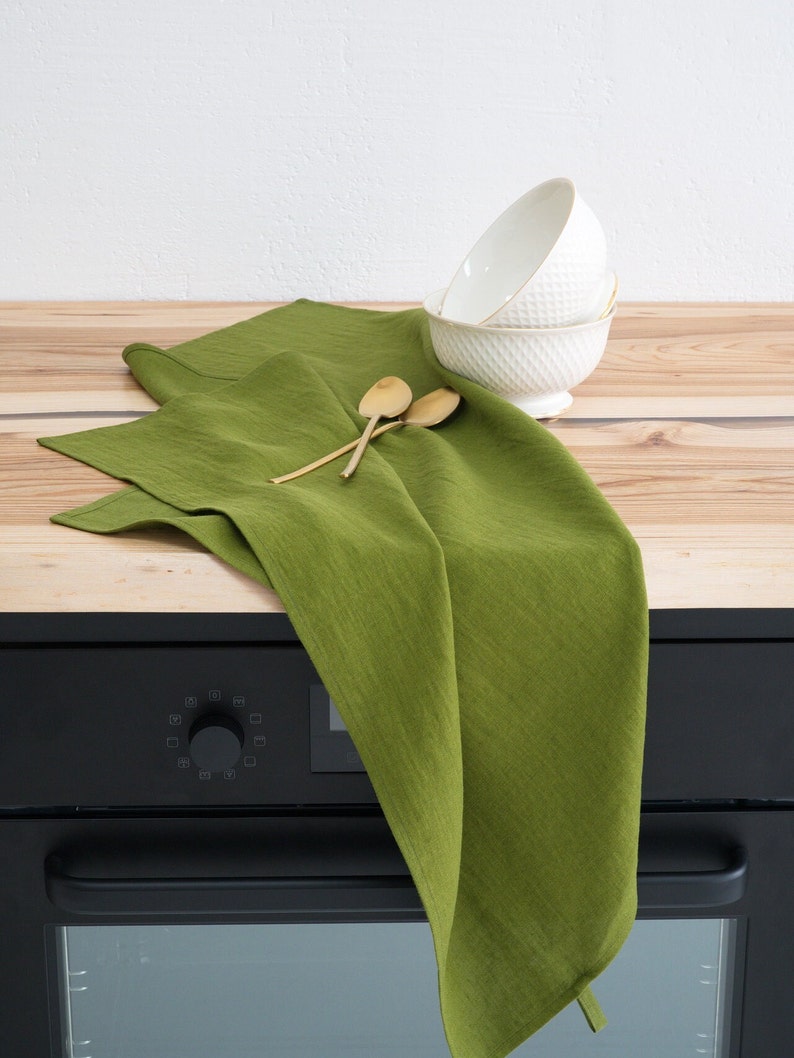 A vibrant green linen towel casually draped over a wooden kitchen countertop, accompanied by a stack of white textured bowls and a pair of wooden spoons, against a white wall.