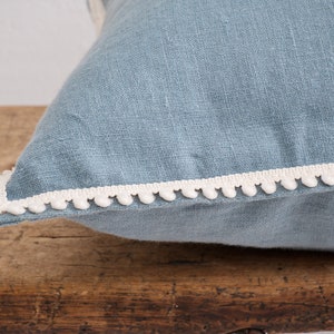 White decorative pom-pom trim on a blue linen fabric pillow resting on a wooden surface.