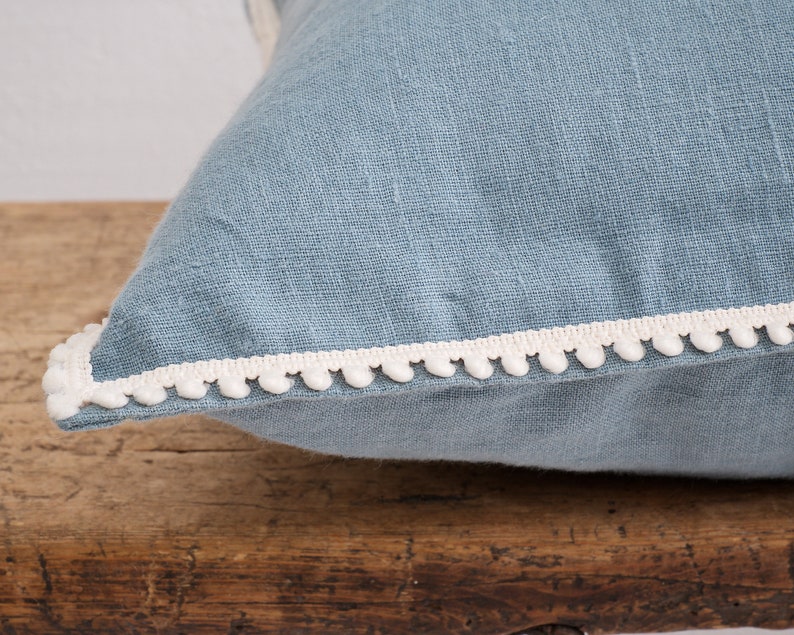 White decorative pom-pom trim on a blue linen fabric pillow resting on a wooden surface.