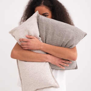 Puede incluir: Una persona sostiene dos almohadas de lino a rayas frente a su cara. Las almohadas son en tonos de marr&oacute;n y blanco.