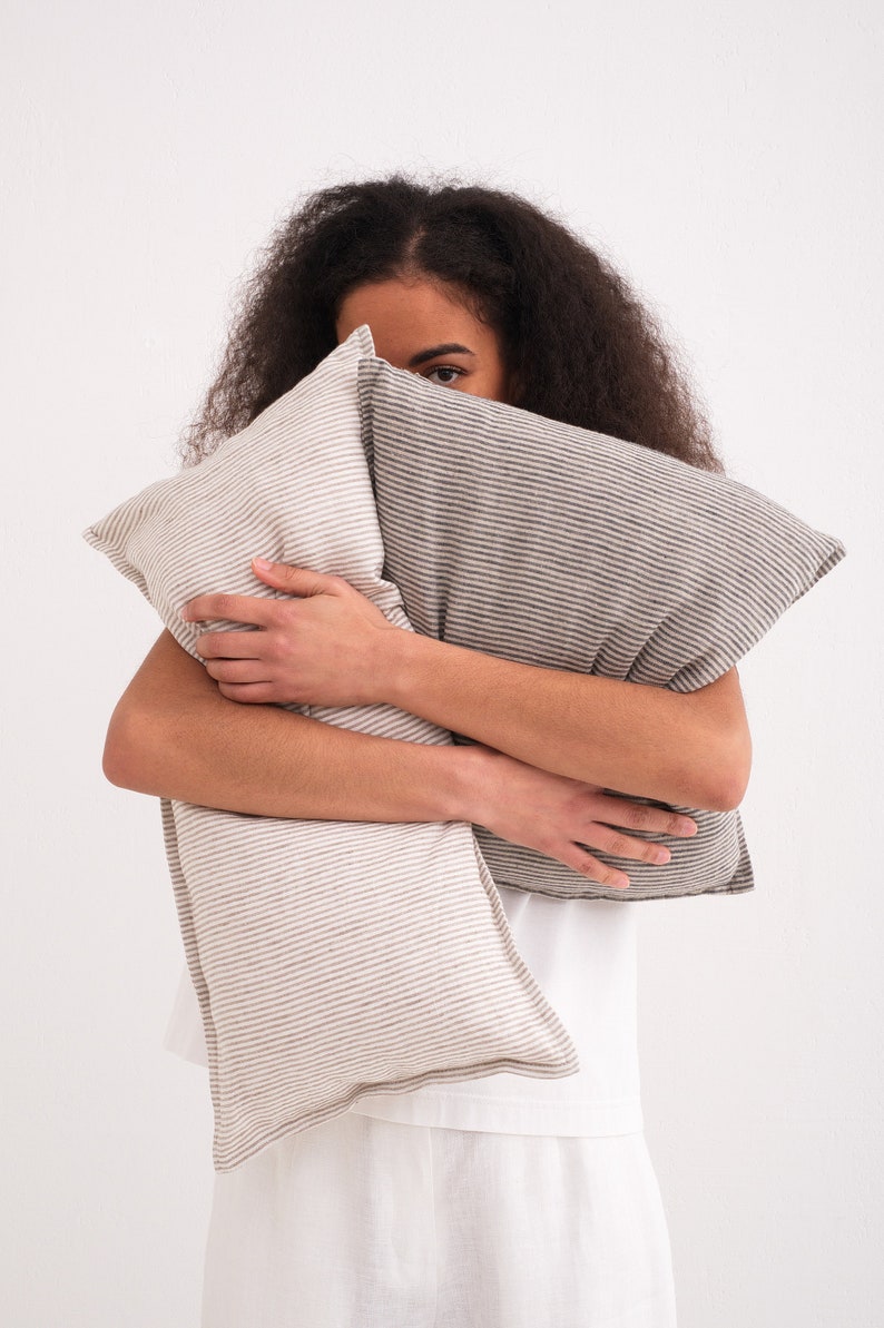 Puede incluir: Una persona sostiene dos almohadas de lino a rayas frente a su cara. Las almohadas son en tonos de marr&oacute;n y blanco.