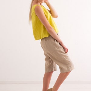 May include: A woman wearing a yellow sleeveless linen top and beige linen shorts. She is standing on a white wooden floor and looking at the camera.