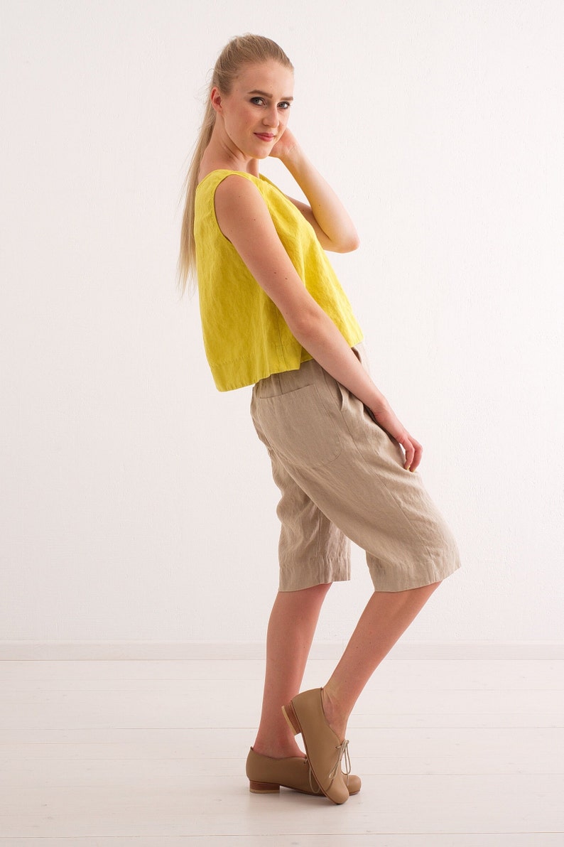 May include: A woman wearing a yellow sleeveless linen top and beige linen shorts. She is standing on a white wooden floor and looking at the camera.