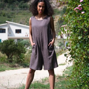 Peut inclure: Une femme portant une combinaison en lin gris sans manches avec des poches. Elle se tient dans une zone herbeuse avec une maison blanche et de la v&eacute;g&eacute;tation verte en arri&egrave;re-plan.