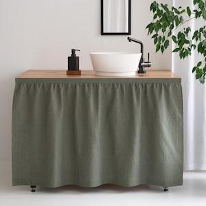 May include: A bathroom vanity with a light wood countertop, a white vessel sink, and a black faucet. A black soap dispenser sits on a wooden coaster. The vanity is covered with a sage green fabric skirt.