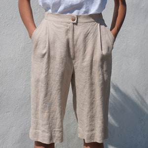 May include: Beige linen shorts with a button and zipper closure. The shorts have pleats in the front and pockets on the sides.