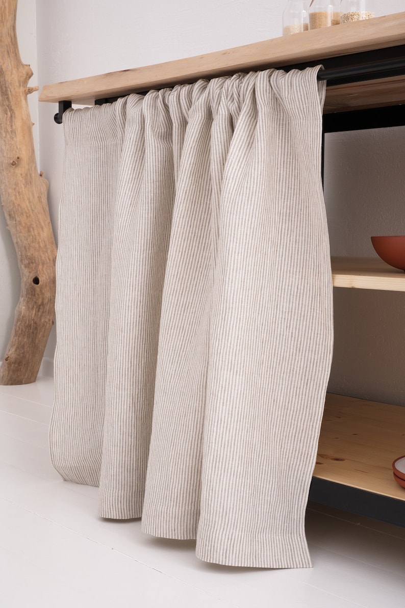 May include: A pair of white linen curtains with thin brown stripes hanging from a wooden rod. The curtains are gathered at the top and hang straight down.