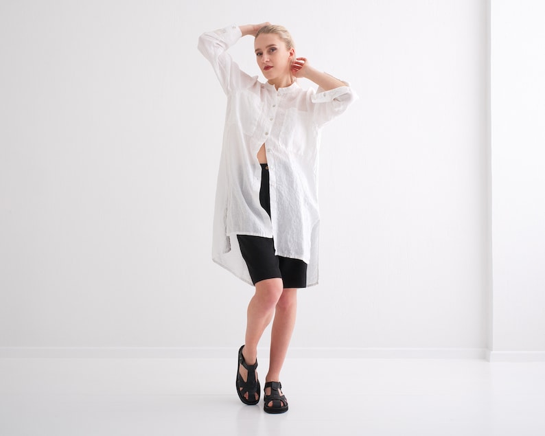 May include: A woman wearing a white linen shirt with a long, flowing design and black shorts. She is standing on a white background and is wearing black sandals.