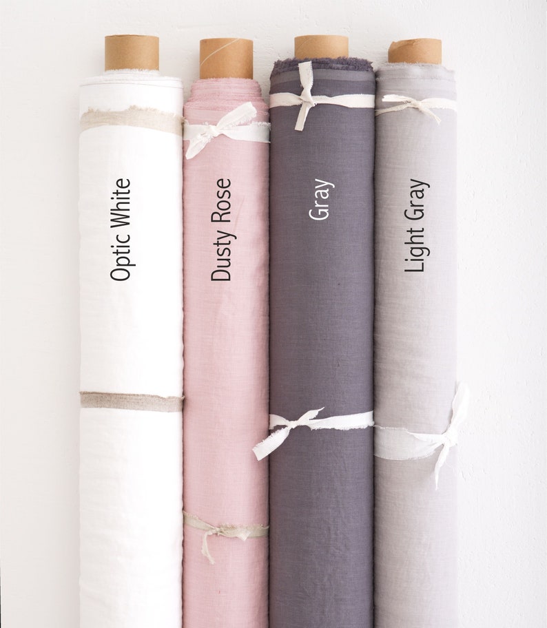 May include: Four rolls of fabric in different colours: optic white, dusty rose, grey, and light grey. Each roll is tied with a white ribbon.
