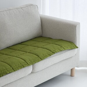 May include: A section of a light gray sofa with a square, olive green cushion. The cushion is quilted with a grid pattern. The sofa has light wooden legs and is positioned near a white curtain.