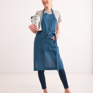 A smiling woman wearing a blue denim apron over a casual outfit holds a metal watering can, paired with skinny jeans and brown sandals, in a bright, airy room.