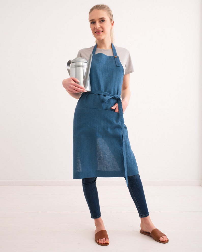 A smiling woman wearing a blue denim apron over a casual outfit holds a metal watering can, paired with skinny jeans and brown sandals, in a bright, airy room.