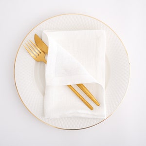 May include: A white linen napkin folded on a white plate with a gold rim. A gold fork and knife are placed on top of the napkin.
