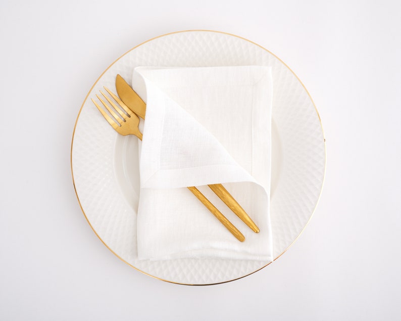 May include: A white linen napkin folded on a white plate with a gold rim. A gold fork and knife are placed on top of the napkin.