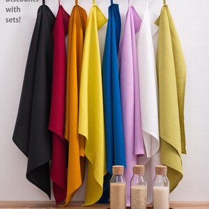 Vividly colored linen towels, ranging from black to bright yellow and cool blue to soft lavender, hang from a modern rack above a wooden table displaying glass bottles with cork lids.