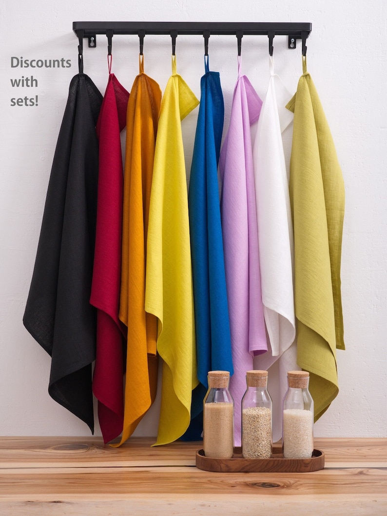 Vividly colored linen towels, ranging from black to bright yellow and cool blue to soft lavender, hang from a modern rack above a wooden table displaying glass bottles with cork lids.