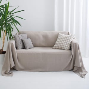 May include: A beige sofa covered with a solid light beige fabric throw. The sofa features three decorative pillows: two smaller grey pillows with visible buttons on the left side and one larger square pillow with a beige and white checkered pattern.