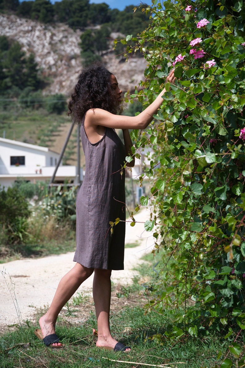 Peut inclure: Une femme v&ecirc;tue d'une robe en lin marron se tient dans un champ et tend la main pour toucher des fleurs roses qui poussent sur une vigne.