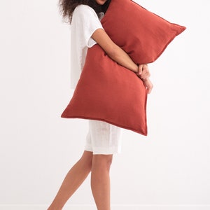 Puede incluir: Una persona con una camisa y pantalones cortos de lino blanco sostiene una almohada de lino de color &oacute;xido.