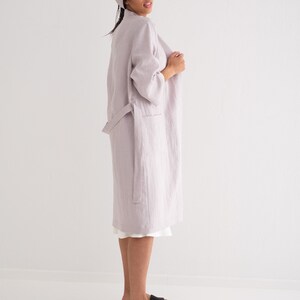 May include: A light purple linen robe with a tie belt and long sleeves. The robe is worn by a person with a towel wrapped around their head. The person is standing on a white surface.