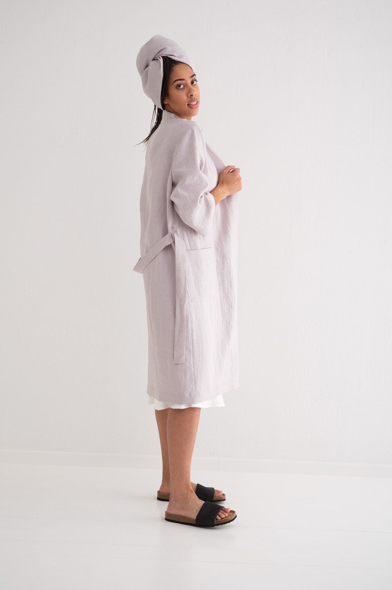May include: A light purple linen robe with a tie belt and long sleeves. The robe is worn by a person with a towel wrapped around their head. The person is standing on a white surface.