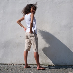 May include: A woman wearing a white sleeveless linen top and beige linen shorts. She is standing in front of a white wall and looking over her shoulder.