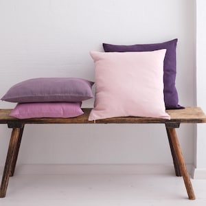 Pode incluir: Uma coleção de quatro almofadas quadradas e retangulares em tons de rosa claro, rosa médio e roxo profundo. As almofadas apresentam uma textura de tecido liso e de cor sólida, dispostas numa pilha e na posição vertical.