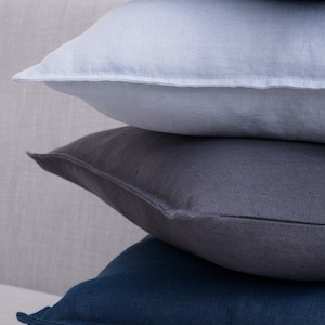 Stacked linen pillow covers in shades of blue, gray, and white on a light gray sofa with wooden armrests, against a white background.