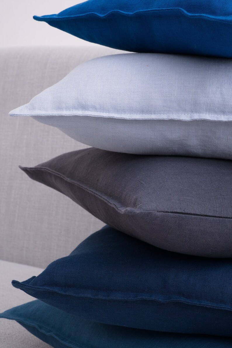 Stacked linen pillow covers in shades of blue, gray, and white on a light gray sofa with wooden armrests, against a white background.