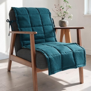 May include: A teal quilted chair cushion is draped over a gray upholstered chair with wooden arms and legs. The cushion has a square pattern and is tied to the chair with black ties. A small wooden side table holds a vase of flowers.