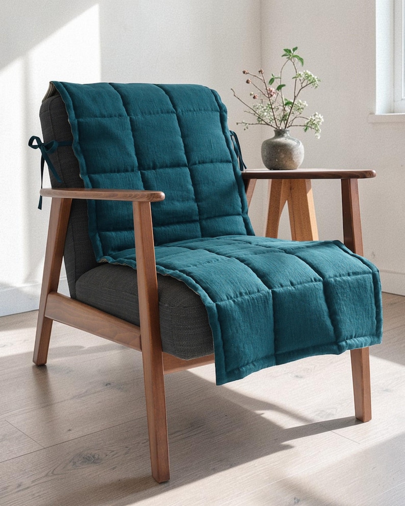 May include: A teal quilted chair cushion is draped over a gray upholstered chair with wooden arms and legs. The cushion has a square pattern and is tied to the chair with black ties. A small wooden side table holds a vase of flowers.