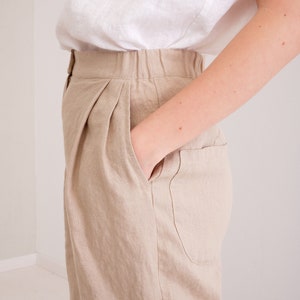 May include: Beige linen pants with a high waist and pleats. The pants have a front pocket and a button closure.