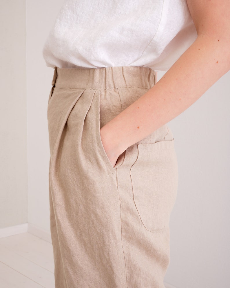 May include: Beige linen pants with a high waist and pleats. The pants have a front pocket and a button closure.