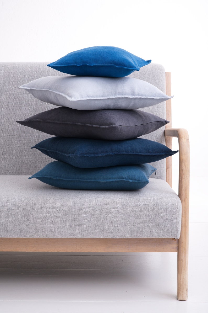 Stacked linen pillow covers in shades of blue, gray, and white on a light gray sofa with wooden armrests, against a white background.