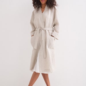 May include: A long, striped linen robe with a sash tie and pockets. The robe is a light beige color with thin brown stripes. The model is wearing the robe and black sandals.