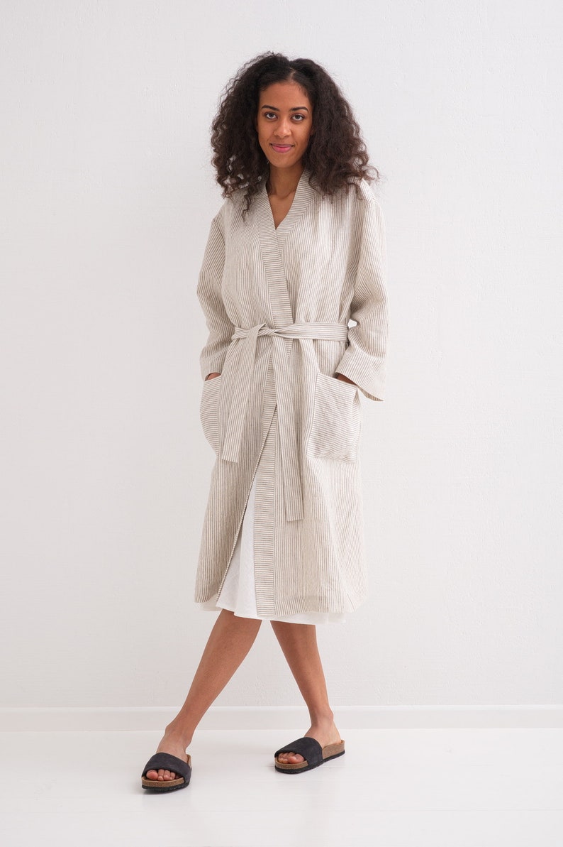 May include: A long, striped linen robe with a sash tie and pockets. The robe is a light beige color with thin brown stripes. The model is wearing the robe and black sandals.