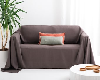May include: A dark grey sofa cover draped over a couch, featuring two rectangular throw pillows in orange and sage green. The fabric has a smooth, matte texture and hangs down to the floor on the sides, covering the entire sofa frame.