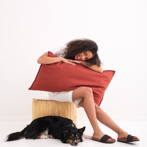 Puede incluir: Una joven con el pelo rizado se sienta en un tronco de madera con una almohada roja. Ella sonr&iacute;e y mira a la c&aacute;mara. Un perro blanco y negro est&aacute; acostado en el suelo frente a ella.