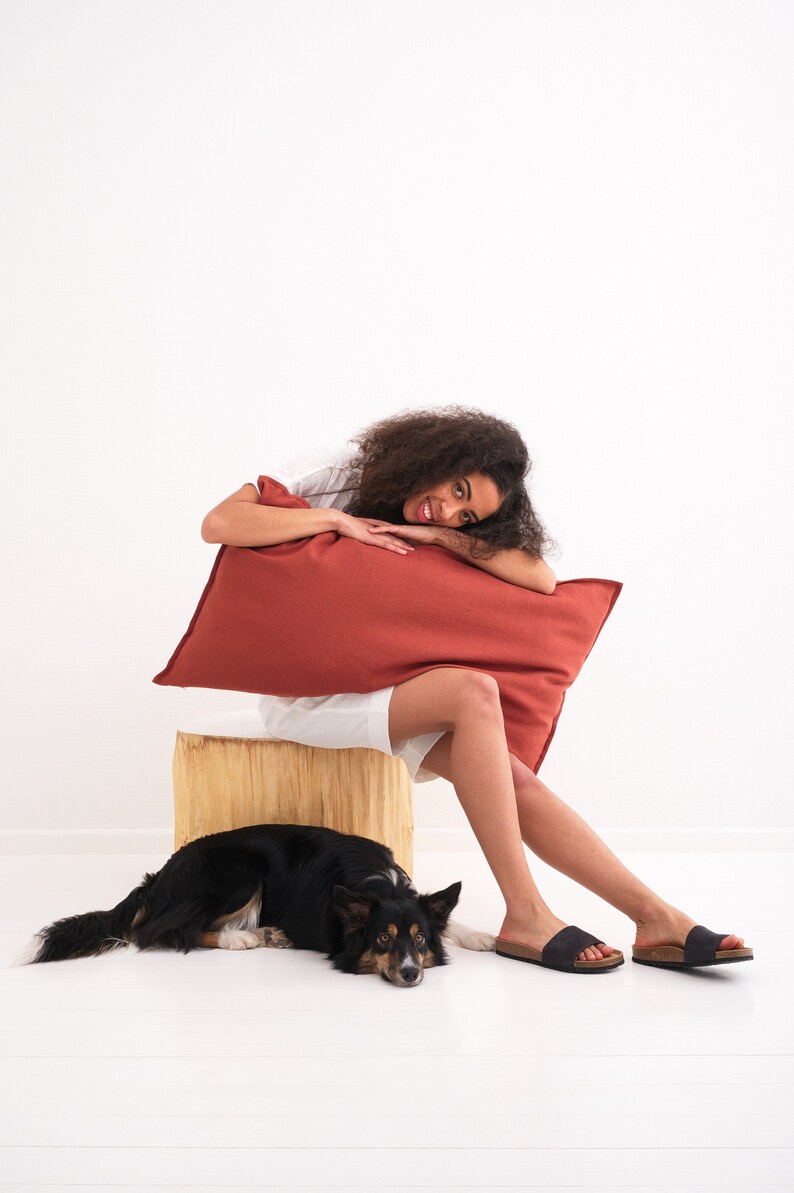 Puede incluir: Una joven con el pelo rizado se sienta en un tronco de madera con una almohada roja. Ella sonr&iacute;e y mira a la c&aacute;mara. Un perro blanco y negro est&aacute; acostado en el suelo frente a ella.