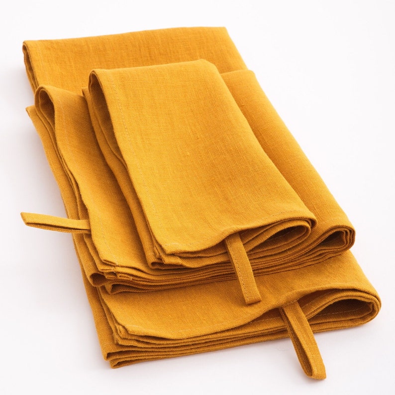 A stack of mustard yellow linen towels neatly folded on a white background, with a soft texture visible on the fabric surface and convenient hanging loops attached to one corner.
