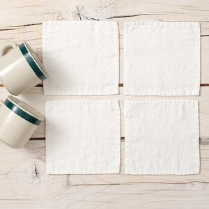 May include: Four white linen coasters on a light wood surface with two beige mugs with green trim.