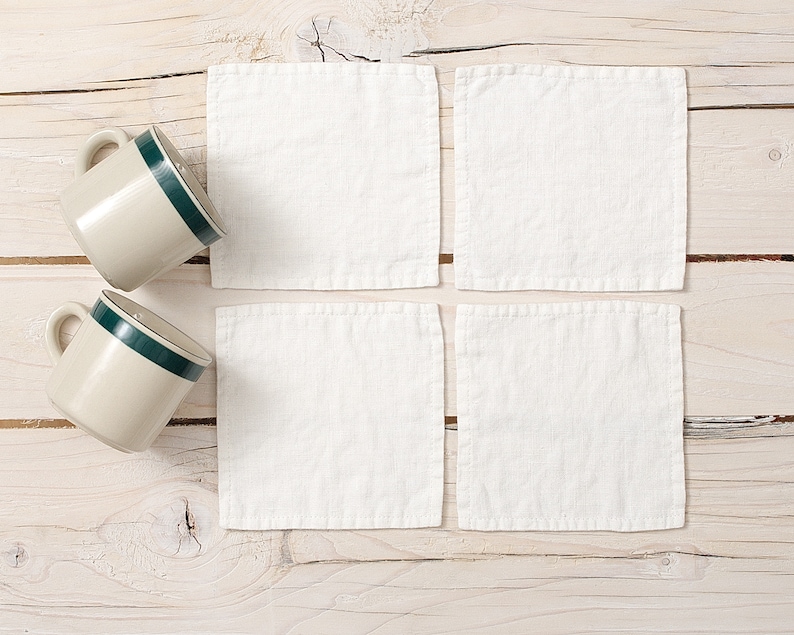 May include: Four white linen coasters on a light wood surface with two beige mugs with green trim.