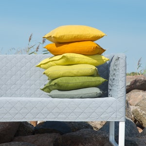 K&ouml;nnte beinhalten: Ein Stapel von f&uuml;nf Kissen in verschiedenen Gelb- und Gr&uuml;nt&ouml;nen, angeordnet auf einem grauen gesteppten Sofa. Das Sofa steht auf einer felsigen Oberfl&auml;che.