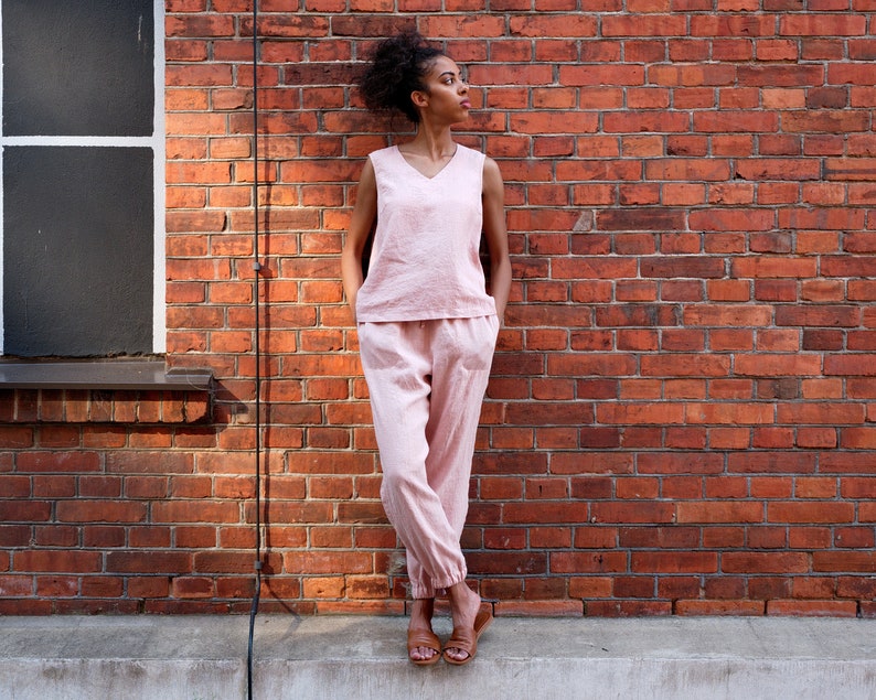 Op de afbeelding: Een vrouw in een roze top zonder mouwen en een bijpassende broek staat voor een bakstenen muur. Ze draagt ​​bruine sandalen.