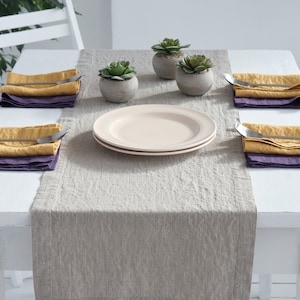 May include: A light gray linen table runner with a white table and four place settings. Each place setting includes a folded yellow linen napkin, a purple linen napkin, and a silver fork. Two small potted succulents are in the center of the table runner.