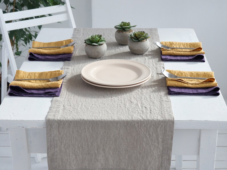 May include: A light gray linen table runner with a white table and four place settings. Each place setting includes a folded yellow linen napkin, a purple linen napkin, and a silver fork. Two small potted succulents are in the center of the table runner.