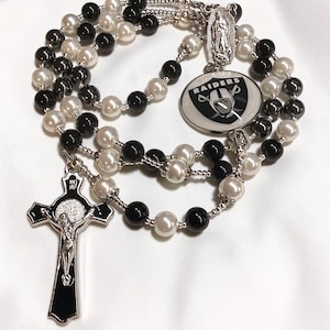 Puede incluir: Un rosario con cuentas alternas negras y blancas tipo perla en una cadena plateada. Incluye un crucifijo plateado con incrustaciones negras, una medalla plateada de la Virgen María y un dije circular que muestra el logotipo del equipo Raiders.