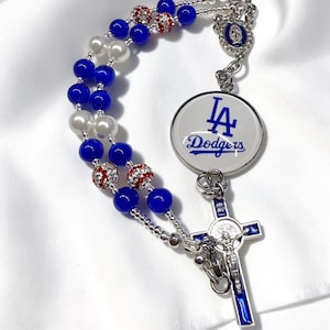 May include: A rosary featuring blue and white beads, red and white baseball-patterned beads, a silver-toned metal crucifix with blue enamel, a circular pendant with the LA Dodgers logo, and a small oval charm depicting a religious figure.